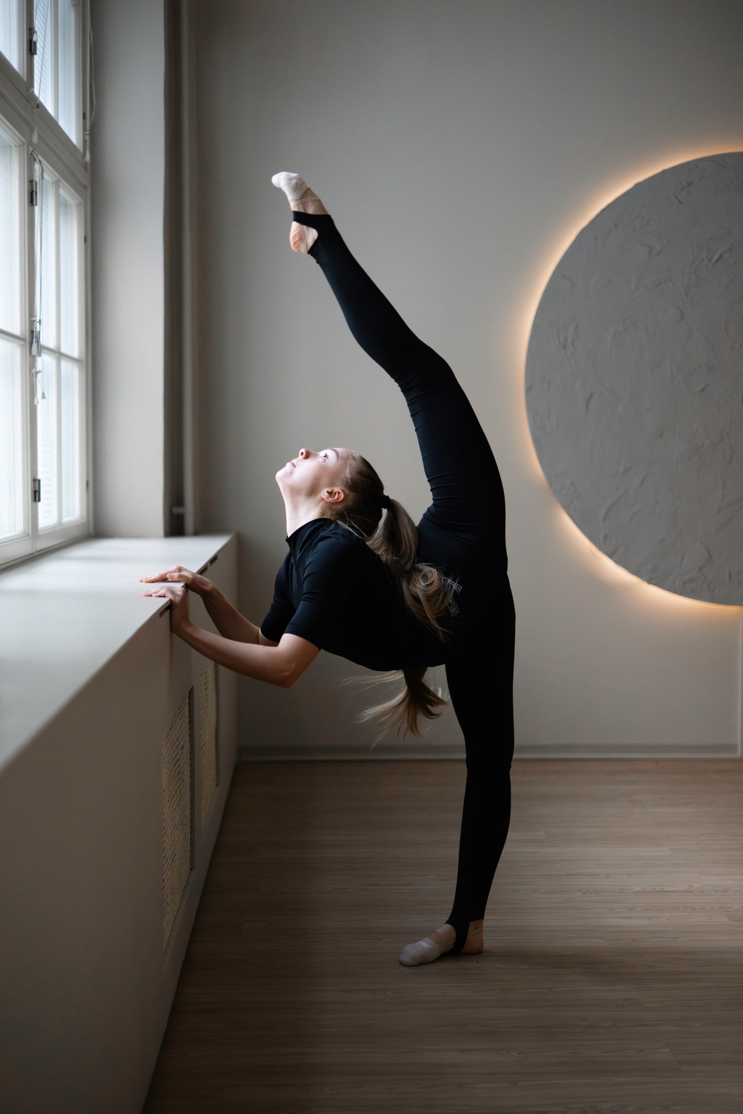 Person in black outfit stretching in a room with a large circular light fixture on the wall.